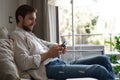 Handsome man in basic t-shirt smiling and holding mobile phone in hands while sitting on couch in living room. Royalty Free Stock Photo