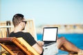 Handsome man on the beach typing on his laptop with white screen for copyspace while sitting on his deck chair Royalty Free Stock Photo