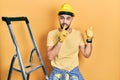 Handsome man with beard by construction stairs wearing hardhat asking to be quiet with finger on lips pointing with hand to the