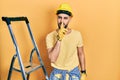 Handsome man with beard by construction stairs wearing hardhat asking to be quiet with finger on lips