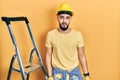 Handsome man with beard by construction stairs wearing hardhat in shock face, looking skeptical and sarcastic, surprised with open Royalty Free Stock Photo