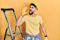 Handsome man with beard by construction stairs wearing hardhat shouting and screaming loud to side with hand on mouth