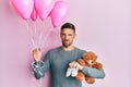 Handsome man with beard expecting a baby girl holding balloons, shoes and teddy bear clueless and confused expression Royalty Free Stock Photo