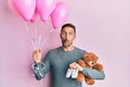 Handsome man with beard expecting a baby girl holding balloons, shoes and teddy bear making fish face with mouth and squinting