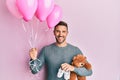 Handsome man with beard expecting a baby girl holding balloons, shoes and teddy bear smiling with a happy and cool smile on face Royalty Free Stock Photo