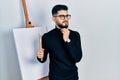 Handsome man with beard holding brushes close to easel stand with hand on chin thinking about question, pensive expression Royalty Free Stock Photo