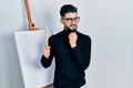 Handsome man with beard holding brushes close to easel stand with hand on chin thinking about question, pensive expression Royalty Free Stock Photo