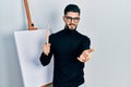 Handsome man with beard holding brushes close to easel stand smiling cheerful offering palm hand giving assistance and acceptance Royalty Free Stock Photo