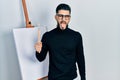 Handsome man with beard holding brushes close to easel stand sticking tongue out happy with funny expression