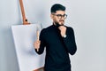 Handsome man with beard holding brushes close to easel stand thinking worried about a question, concerned and nervous with hand on Royalty Free Stock Photo