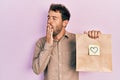 Handsome man with beard holding delivery paper bag with heart reminder bored yawning tired covering mouth with hand Royalty Free Stock Photo