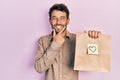 Handsome man with beard holding delivery paper bag with heart reminder looking confident at the camera smiling with crossed arms Royalty Free Stock Photo