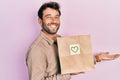 Handsome man with beard holding delivery paper bag with heart reminder pointing aside with hands open palms showing copy space, Royalty Free Stock Photo