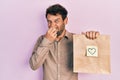 Handsome man with beard holding delivery paper bag with heart reminder smelling something stinky and disgusting, intolerable