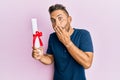 Handsome man with beard holding graduate degree diploma covering mouth with hand, shocked and afraid for mistake