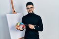 Handsome man with beard holding palette and brush close to easel stand smiling with a happy and cool smile on face Royalty Free Stock Photo