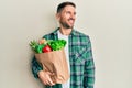 Handsome man with beard holding paper bag with groceries looking away to side with smile on face, natural expression