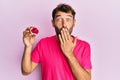 Handsome man with beard holding pink geode precious gemstone covering mouth with hand, shocked and afraid for mistake