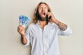 Handsome man with beard and long hair talking on the phone holding 100 brazilian reals angry and mad screaming frustrated and Royalty Free Stock Photo