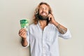 Handsome man with beard and long hair talking on the phone holding 50 shekels angry and mad screaming frustrated and furious, Royalty Free Stock Photo