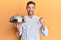 Handsome man with beard wearing chef uniform holding silver tray pointing thumb up to the side smiling happy with open mouth Royalty Free Stock Photo