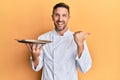 Handsome man with beard wearing chef uniform holding silver tray pointing thumb up to the side smiling happy with open mouth Royalty Free Stock Photo