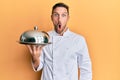 Handsome man with beard wearing chef uniform holding silver tray scared and amazed with open mouth for surprise, disbelief face Royalty Free Stock Photo