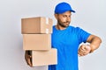 Handsome man with beard wearing courier uniform holding delivery packages checking the time on wrist watch, relaxed and confident Royalty Free Stock Photo