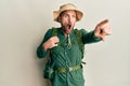 Handsome man with beard wearing explorer hat and backpack pointing with finger surprised ahead, open mouth amazed expression,