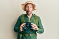 Handsome man with beard wearing explorer hat looking through binoculars making fish face with mouth and squinting eyes, crazy and Royalty Free Stock Photo