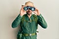 Handsome man with beard wearing explorer hat looking through binoculars relaxed with serious expression on face Royalty Free Stock Photo