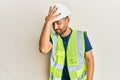 Handsome man with beard wearing safety helmet and reflective jacket surprised with hand on head for mistake, remember error