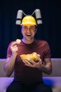 Handsome man with beer helmet on the head watching TV and eating potato chips on the couch at home. Young male sits on a Royalty Free Stock Photo