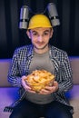 Handsome man with beer helmet on the head watching TV and eating potato chips on the couch at home. Young male sits on a Royalty Free Stock Photo