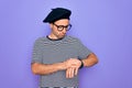 Handsome man with blue eyes wearing striped t-shirt and french beret over purple background Checking the time on wrist watch, Royalty Free Stock Photo