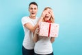 A handsome man closes the eyes of his woman, making her a surprise, and gives her a gift against a blue background