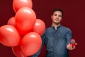 Handsome man with coral balloons and red box with engagement ring  over dark red background. Royalty Free Stock Photo