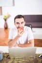 Handsome man eating apple and working with laptop at kitchen at home. Royalty Free Stock Photo