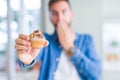 Handsome man eating chocolate chips muffin cover mouth with hand shocked with shame for mistake, expression of fear, scared in