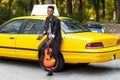 Handsome man with guitar in casual clothes near yellow taxi, sitting near door, posing confident. Horizontal view.