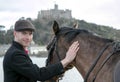 Handsome man, Male Horse Rider pets his horse on beach, wearing traditional flat cap, white trousers, red polo shirt Royalty Free Stock Photo