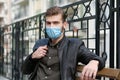 Handsome man in medical protective face mask sits on bench and looks at camera