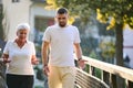 A handsome man and an older woman share a serene walk in nature, crossing a beautiful bridge against the backdrop of a Royalty Free Stock Photo