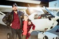 Handsome man in pilot jacket stands with kid brother near hangar with single-engine airplane