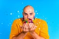 handsome man with raindow in beard and yellow sweater blowing glitter confetti in blue studio baclground