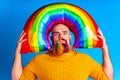 handsome man with raindow colors in beard and yellow sweater holding balloon inflatable above of his head in blue studio