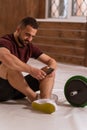 Handsome man sitting on a floor with smartphone in his hands and black and green tone fitness barbell, equipment for