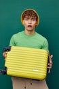 a handsome man stands on a green background, dressed in a yellow panama hat, holding a bright travel suitcase in his Royalty Free Stock Photo