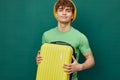 a handsome man stands on a green background, dressed in a yellow panama hat, holding a bright travel suitcase in his Royalty Free Stock Photo