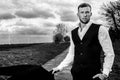 Handsome man in suit and waistcoat standing with fields and stormy sky behind him looking at camera.